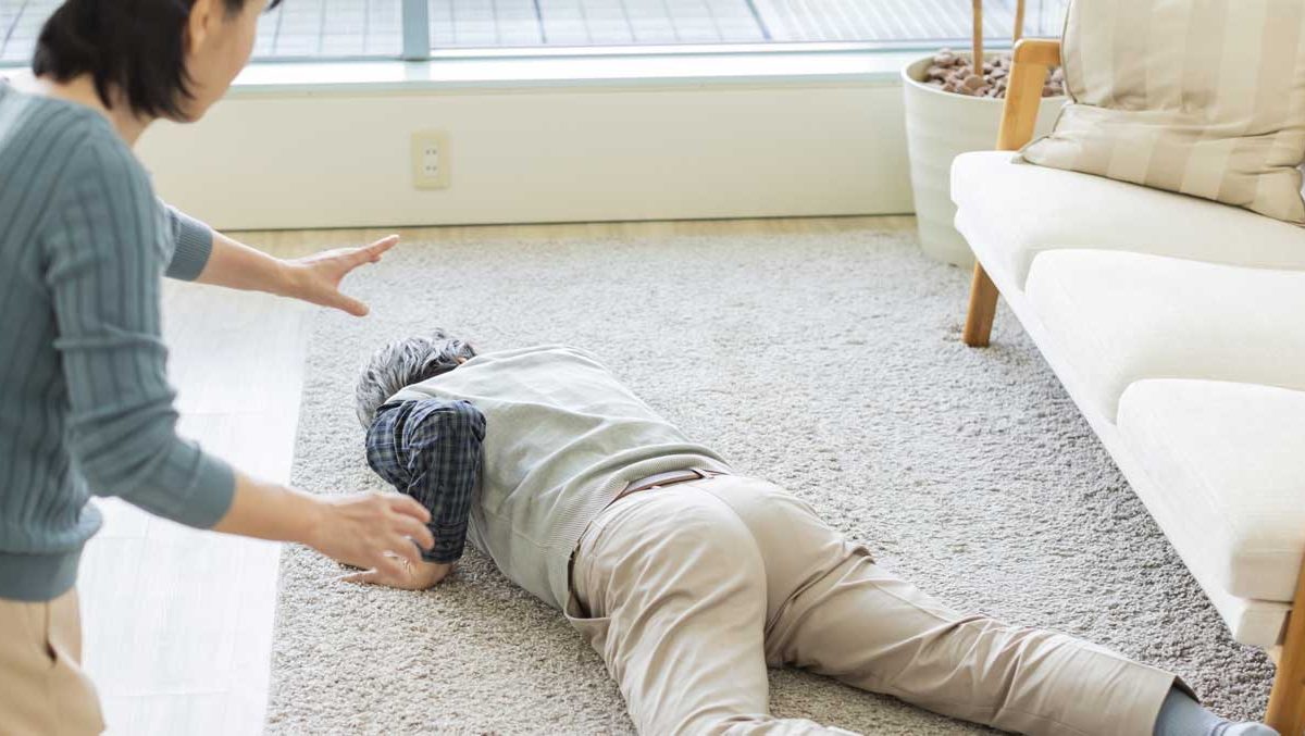 A senior citizen lies facedown on a rug in medical distress, as a woman approaches to help.